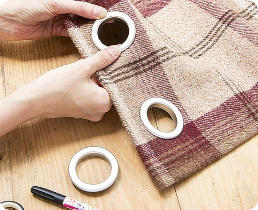 Hands insert eyelets into plaid fabric on a wooden work surface.